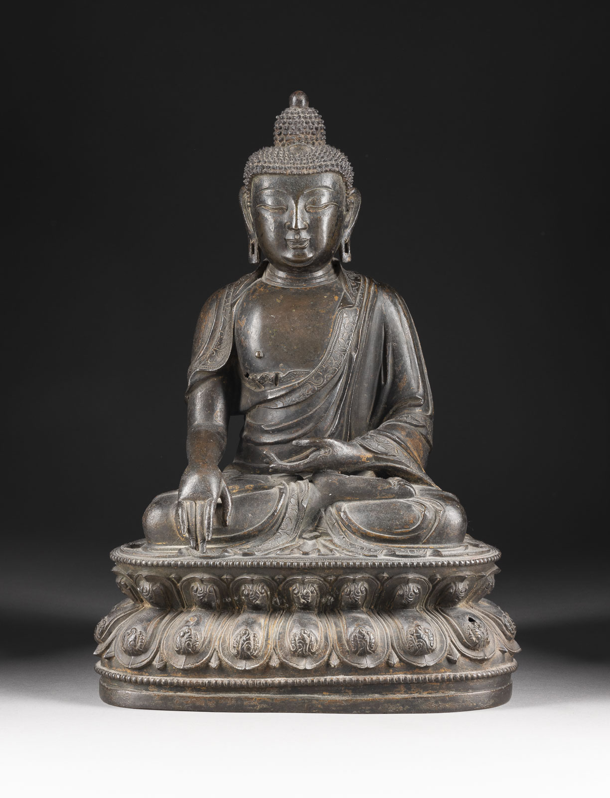 A detailed bronze statue of a seated Buddha on an elaborately decorated lotus throne. The Buddha wears a traditional robe and a crown, sitting in a meditation posture with his right arm raised and his hand resting flat on his knee. The background is uniformly dark, highlighting the statue.