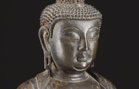Nahaufnahme einer antiken Buddha-Statue aus Metall, die eine ruhige und meditative Ausdruckskraft zeigt. Die Statue ist detailreich gestaltet, mit sichtbaren Mustern auf dem Gewand und einer fein strukturierten Haarkrone. Der Hintergrund ist einheitlich dunkel, um die Figur hervorzuheben.