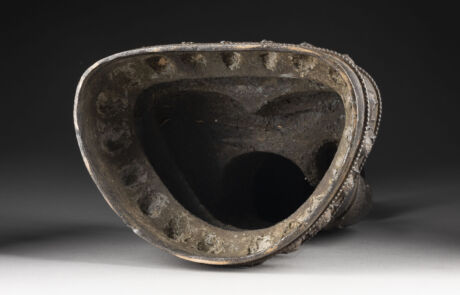 Dieses Bild zeigt ein antikes, dunkles Metallgefäß mit einer ungewöhnlichen, abgerundeten und eingedellten Form, welches eine starke Patina und Verschleißspuren aufweist. Das Objekt steht auf einer hellen Oberfläche vor einem dunkelgrauen Hintergrund. Es scheint Teil einer Auktion von historischen oder archäologischen Funden zu sein.