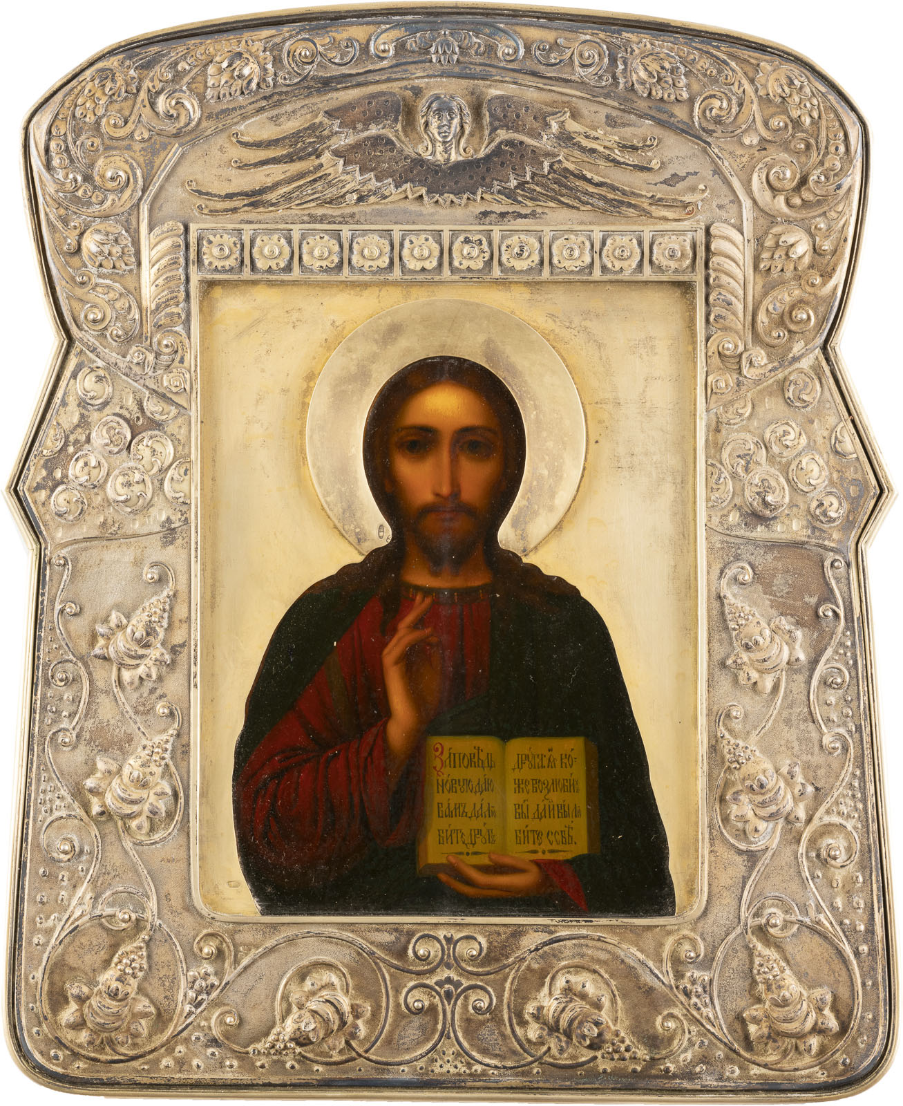The image shows a religious icon depicting a central portrait of Jesus Christ with a gentle, serious gaze, surrounded by a halo. He wears a red robe and holds an open book with Cyrillic script in his left hand. The icon is set in a lavishly decorated silver frame decorated with floral patterns.