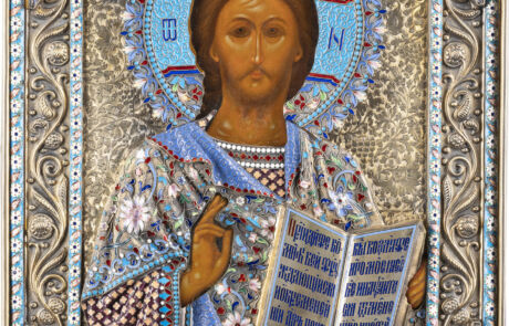 Detailreiches, orthodoxes Ikonengemälde, das Jesus Christus darstellt, umgeben von einem aufwendigen, silbernen und blau verzierten Heiligenschein. Jesus trägt eine bestickte blaue und weiße Gewandung und hält ein aufgeschlagenes Buch mit kyrillischer Schrift. Sein Gesichtsausdruck ist ernst und introspektiv.