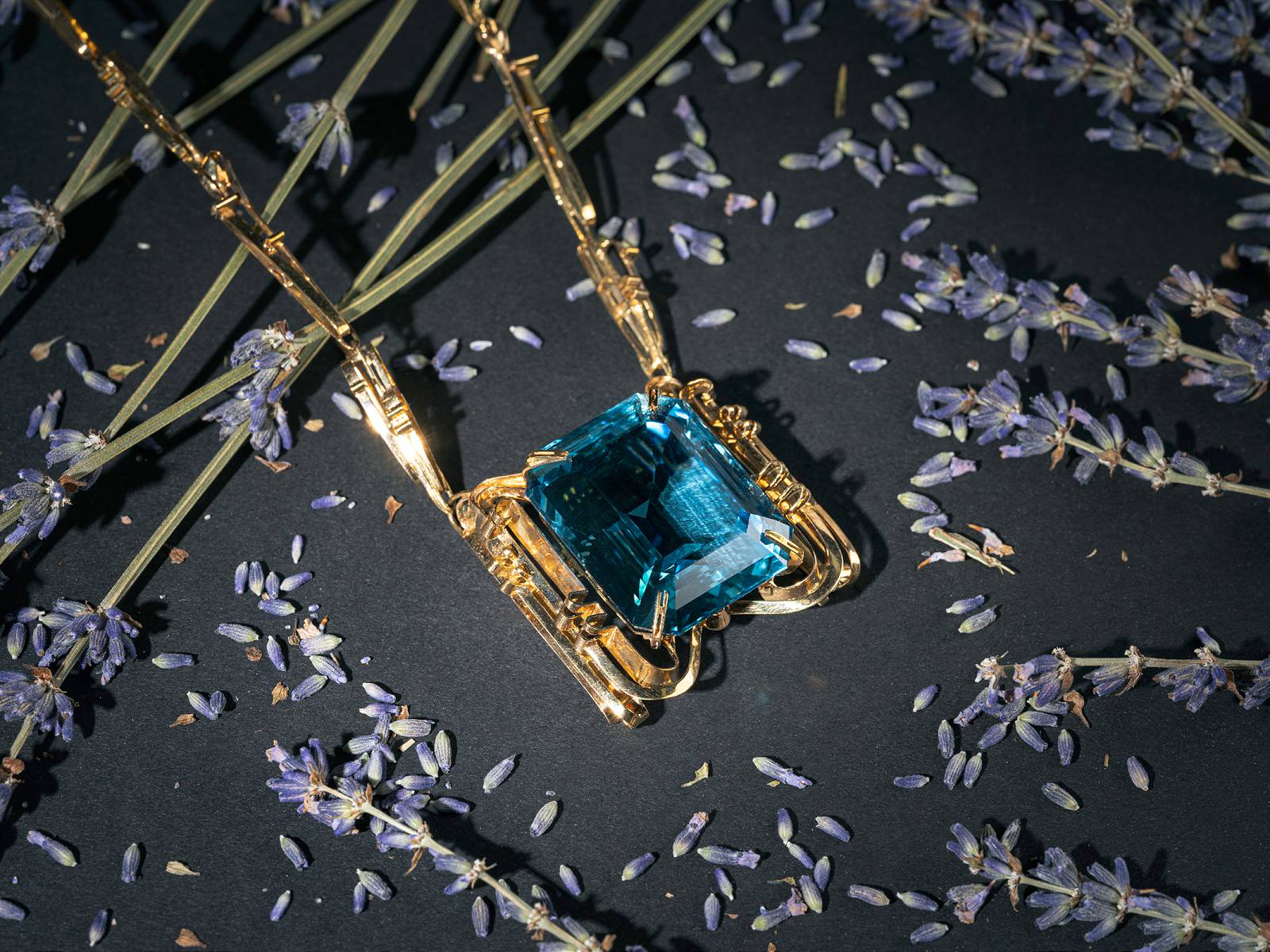 Großer blauer Edelstein in goldener Fassung auf dunklem Hintergrund umgeben von Lavendelzweigen und Blüten.