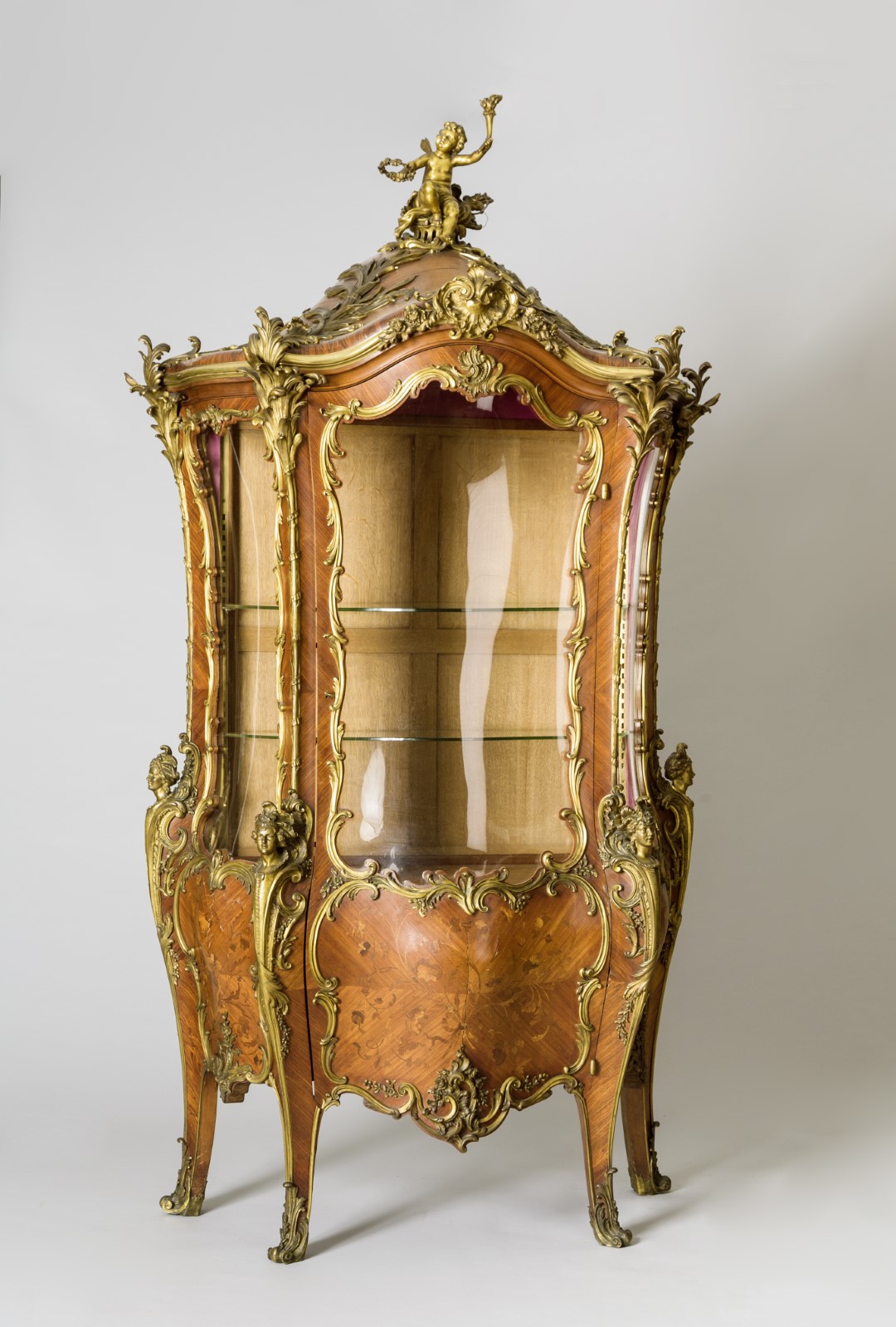Opulent, Rococo-style wooden display cabinet, richly decorated with gilded ornaments. It features curved legs and a vaulted roof topped with a figure. The front and sides are fitted with cut glass panels, allowing glimpses into the interior with two glass shelves.