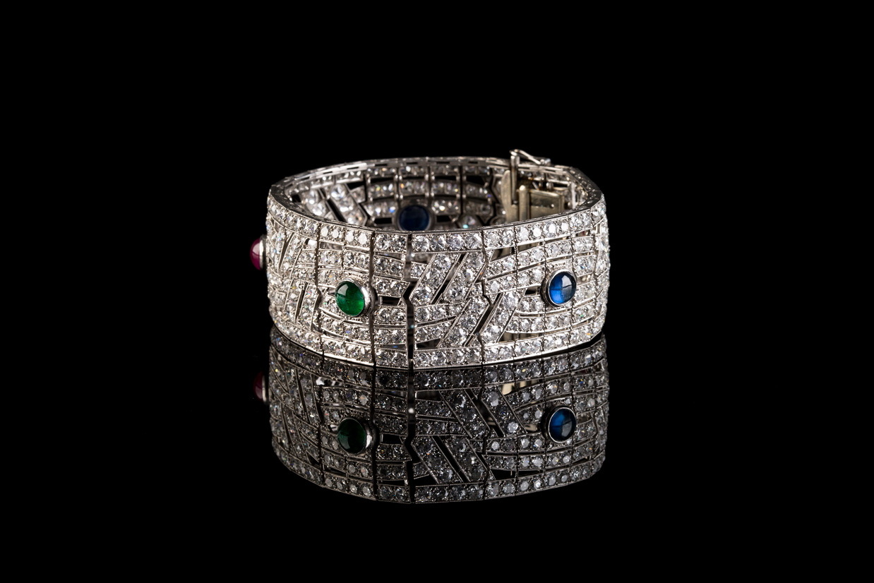 The image shows a luxurious, wide bracelet, richly encrusted with diamonds, presented against a black background. The bracelet features a complex, geometric design and is adorned with several colored gemstones, including green and blue.