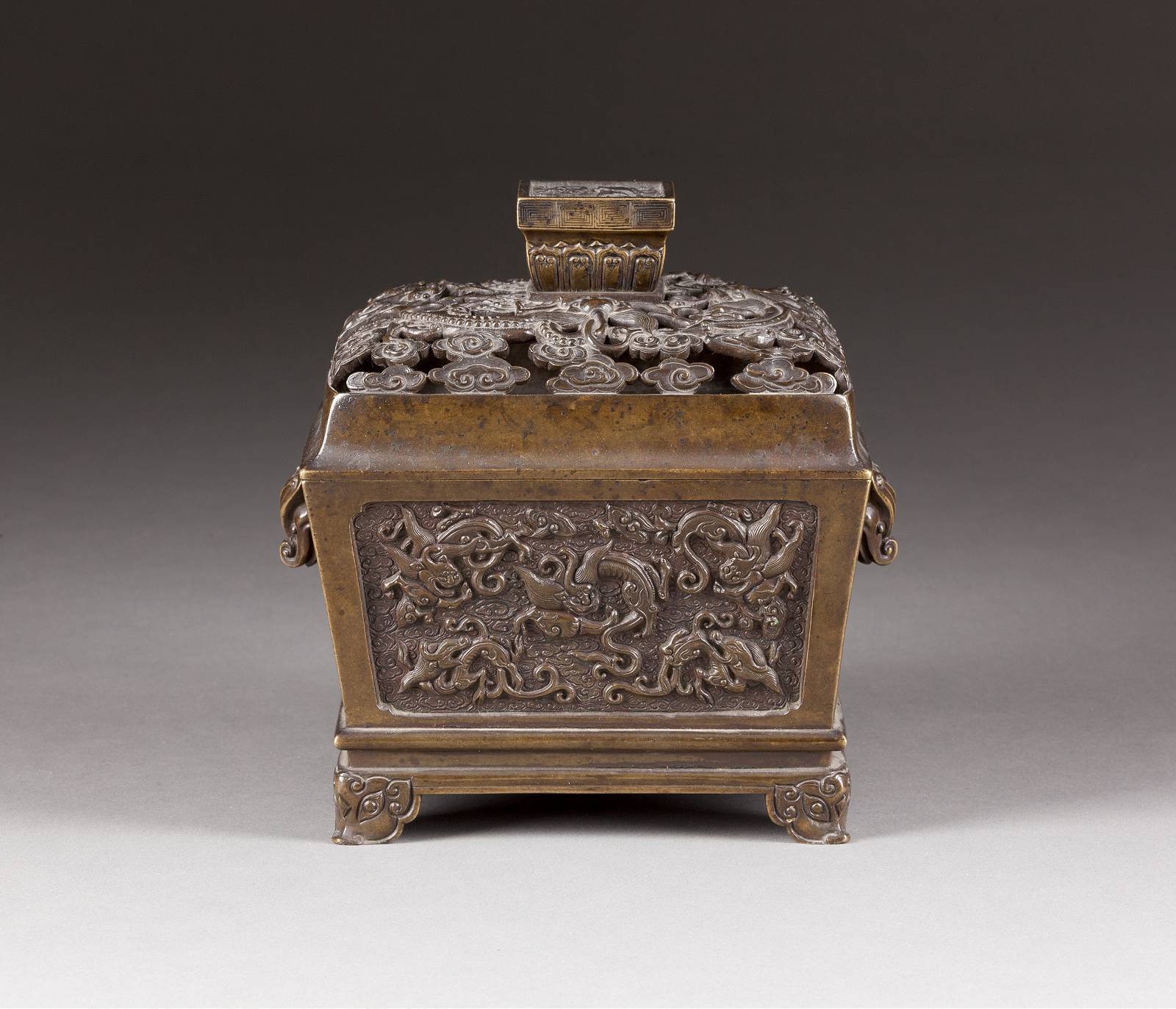 An antique, rectangular metal container on a small pedestal, probably made of bronze, with complex, engraved decorations on all sides. The lid of the container is crowned with finely crafted floral designs, terminating in a prominent, rectangular handle.