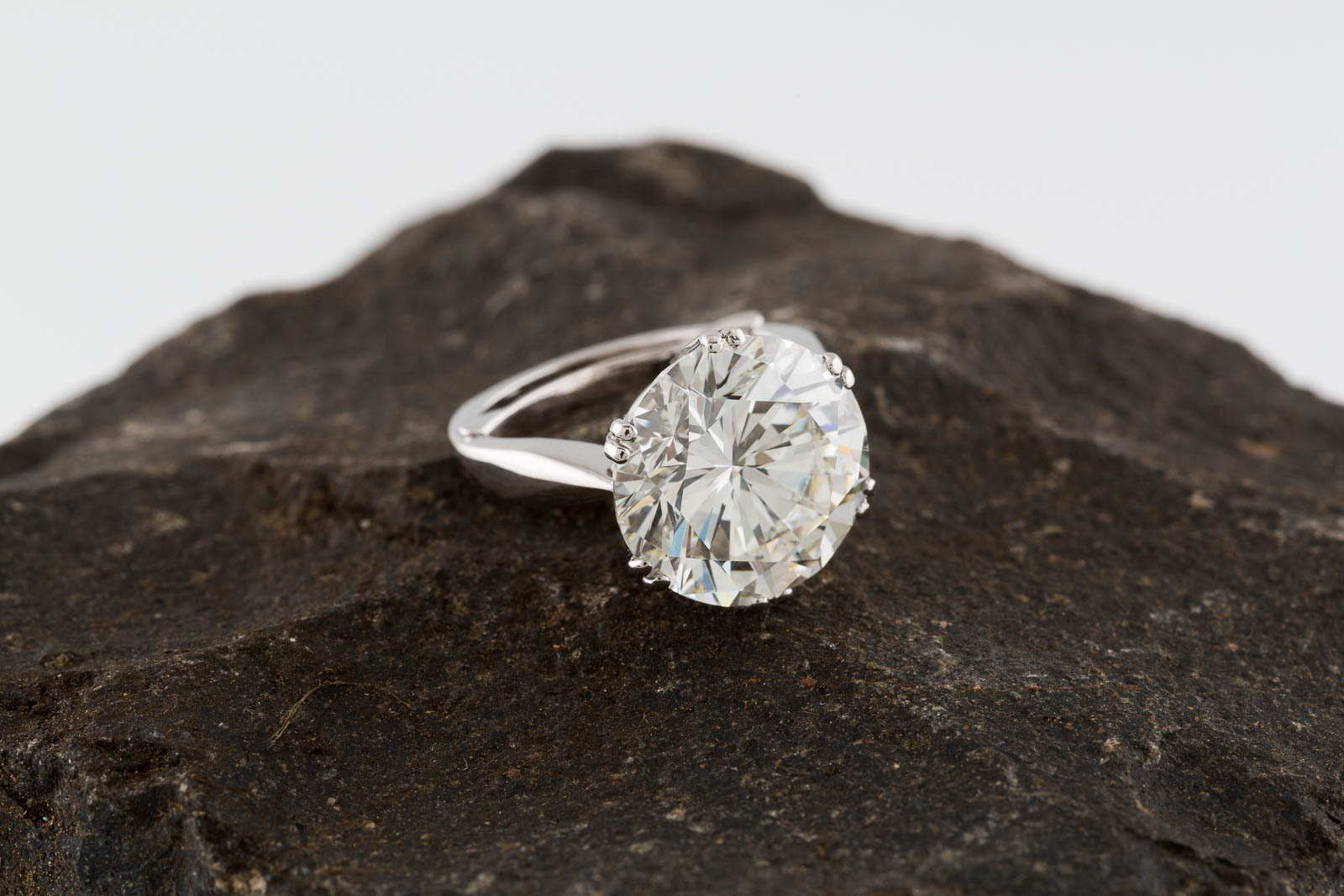 A lustrous diamond ring set on a dark, rough rock, featuring a large, brightly sparkling solitaire diamond in a silver setting. The diamond is round-cut, and the metal of the ring shines with its polish.