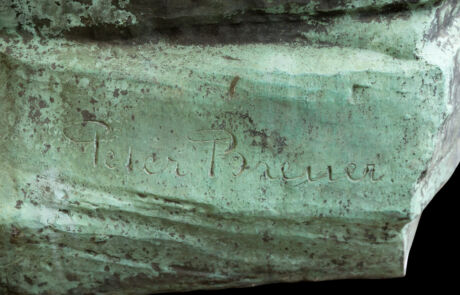 Nahaufnahme eines kunstvollen, grün patinierten Metallfragments, vermutlich ein Teil einer Skulptur oder Plakette. Auf der leicht verwitterten Oberfläche ist sichtbar und in geschwungener Schrift eingraviert: "Peter Breuer". Das Stück suggeriert antike oder historische Bedeutung, typisch für Auktionsgegenstände.