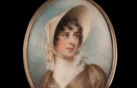 WILLIAM WOOD 1769 Suffolk – 1810 London FEINE PORTRÄTMINIATUR EINER DAME MIT HUT This image shows a portrait of a woman from an earlier era, set in an oval, metallic frame. The person depicted wears a large, light-colored cap and a dark brown dress with a plunging neckline and white lace collar. Her gaze appears thoughtful, and she has slight blushes on her cheeks.