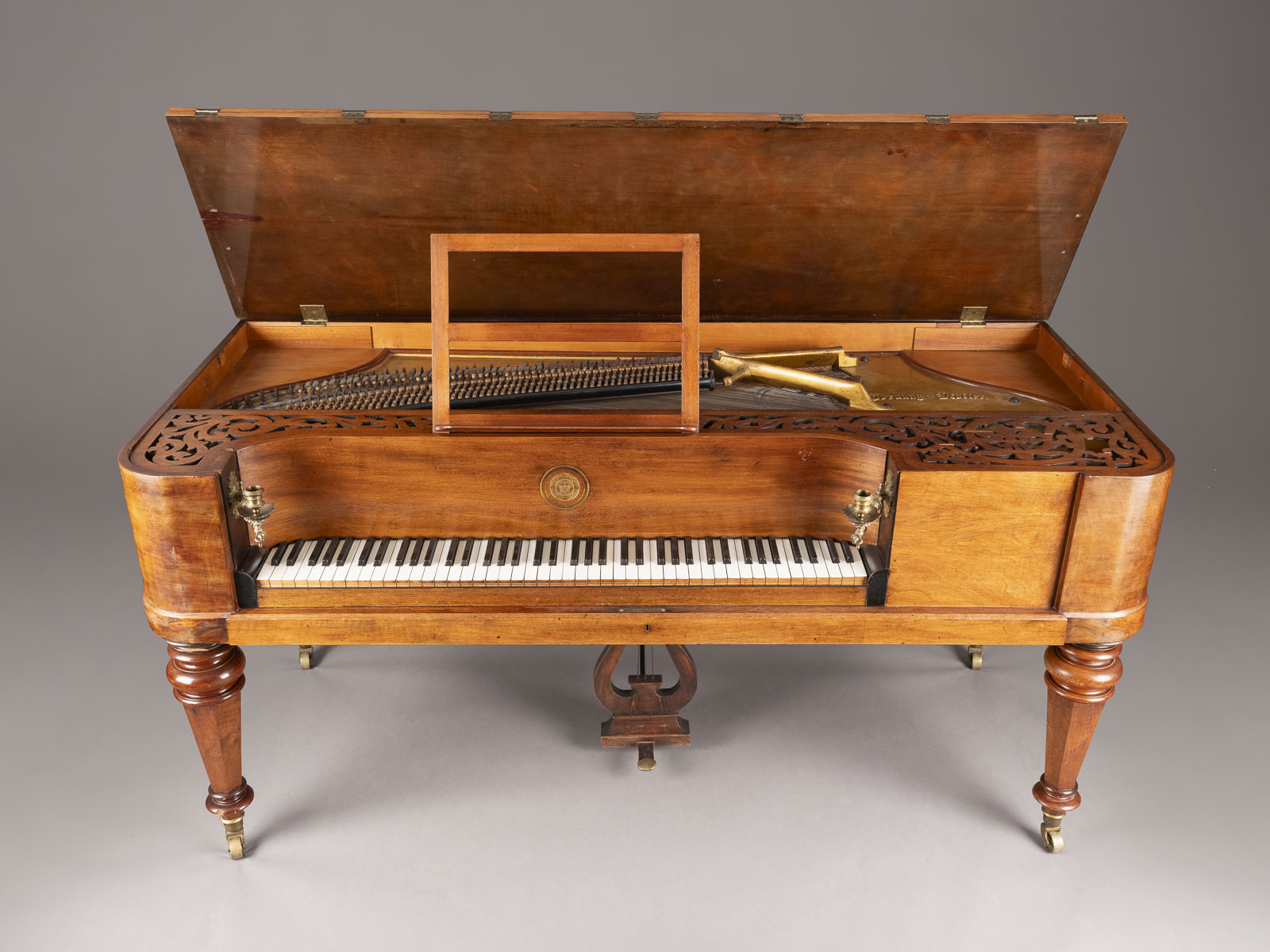 View of an open, antique grand piano on tall, turned legs with casters. The polished wooden instrument features intricate carving on the front panel. The keyboard is fully visible, and the lid is raised, revealing the internal mechanism and strings.