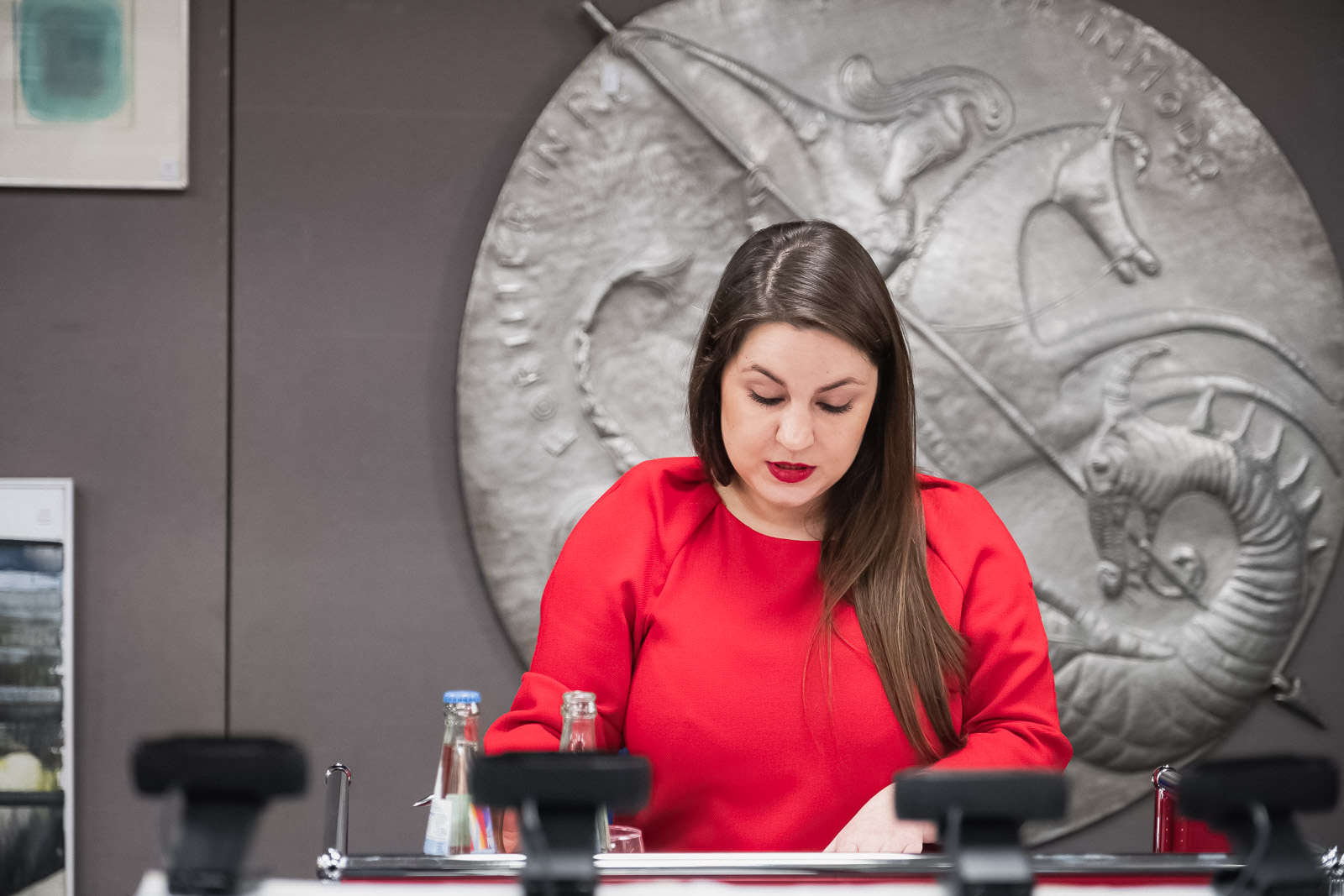 Eine Frau in einem roten Kleid konzentriert sich auf das Lesen von Dokumenten bei einer Kunstauktion. Hinter ihr ist eine große, runde Metallplatte mit erhabenen Reliefs von mythologischen Kreaturen und Inschriften an einer grauen Wand angebracht. Im Vordergrund sind undeutlich Mikrofone und Wasserflaschen auf einem Tisch zu erkennen.