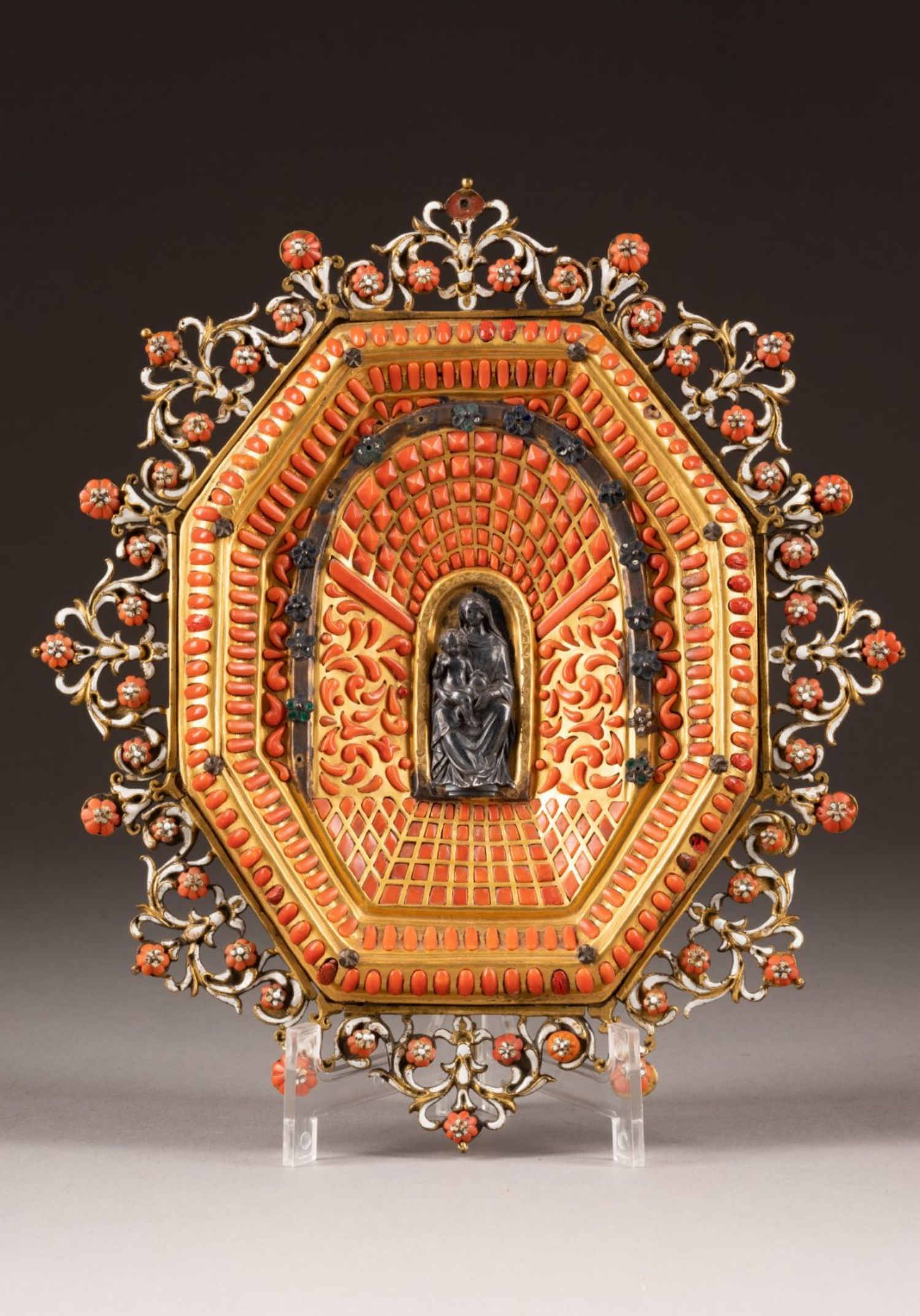 Detailliertes, achteckiges Kunstobjekt, vermutlich eine Art Reliquienkapsel oder Altar, mit einem zentralen, schwarzen Medaillon, das eine Reliefdarstellung einer religiösen Figur zeigt. Das Objekt ist reich verziert mit goldenen und roten Ornamenten sowie floralen Motiven, die mit Perlmutt-ähnlichen Akzenten besetzt sind.