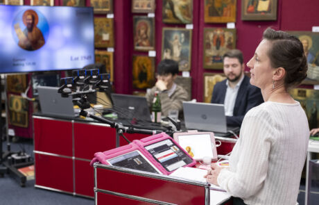 Auktionatorin leitet Kunstauktion in Saal voller historischer Kunstwerke, Personen bieten per Computer.