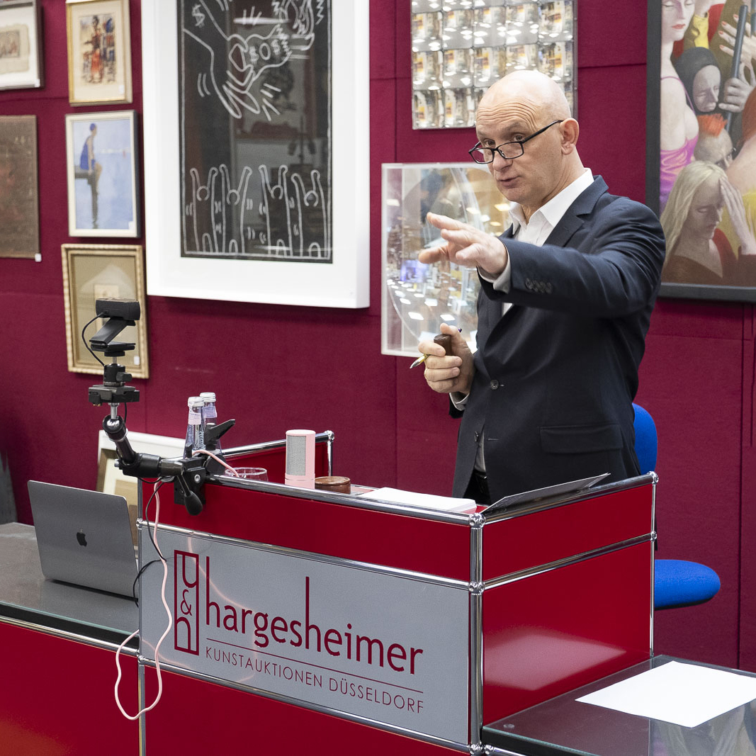 Auktionator bei einer Kunstauktion im Auktionshaus Hargesheimer in Düsseldorf, gestikuliert hinter einem Pult, umgeben von verschiedenen Kunstwerken.
